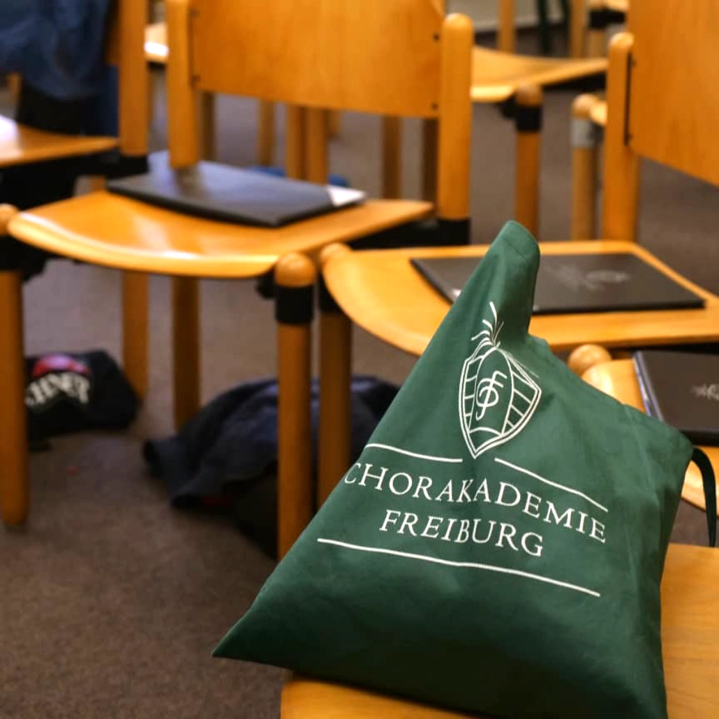 In einem Probensaal stehen Stühle. Darauf liegen Notenmappen und Taschen mit Chorakademie Freiburg Logo