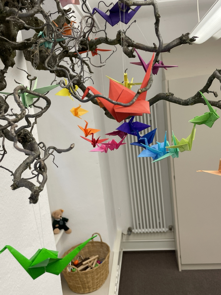 Kursraum Mobile Kranich Viele bunte Kraniche aus Origami gefaltet flattern um einen Ast herum