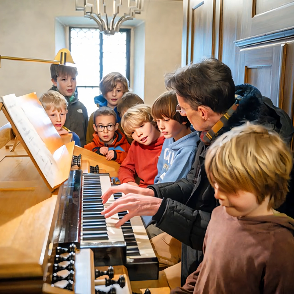 Einsteigerchor der Chorakademie Freiburg Kinder im Grundschulalter stehen an einer Orgel in einer Kirche. An der Orgel sitzt Pr. Böhmann und erklärt das Instrument