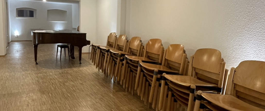 Probensaal Chorakademie Freiburg In einem großen Probensaal stehen viele Stühle aufeineander gestapelt am Rand. In der Mitte steht ein brauner Klavieflügel